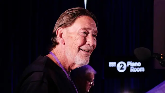 Chris Rea smiles as he stands in front of a microphone in the BBC Radio 2 Piano Room