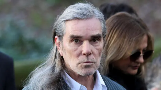Ian Brown wearing a pinstripe suit and white shirt, he has long hair partly tucked behind his ears.
