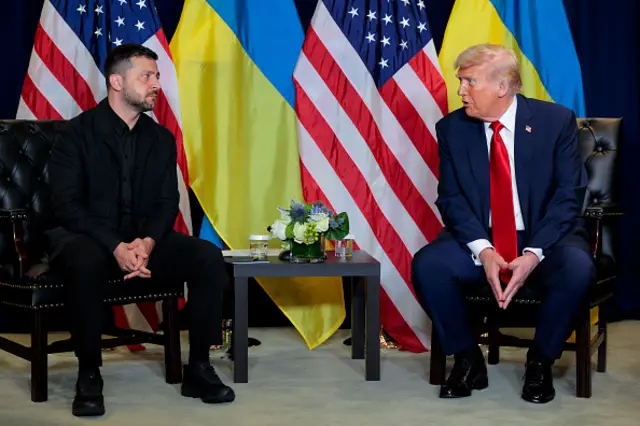Volodymyr Zelensky and Donald Trump sit next to each other on black chairs, with a small black table in the middle, and US and Ukrainian flags behind them.