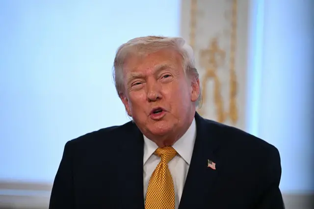 Donald Trump in black suit, white shirt and golden tie mid-speech