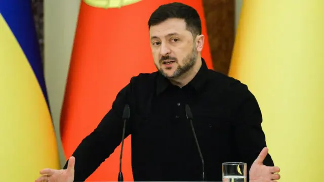 A black-clad Volodymyr Zelensky speaks in front of microphones with Ukrainian and Portuguese flags behind him.