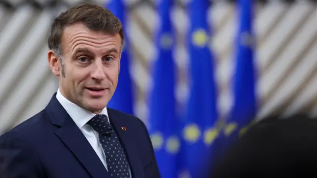 Emmanuel Macron in a dark suit against a blurred blue and grey background.