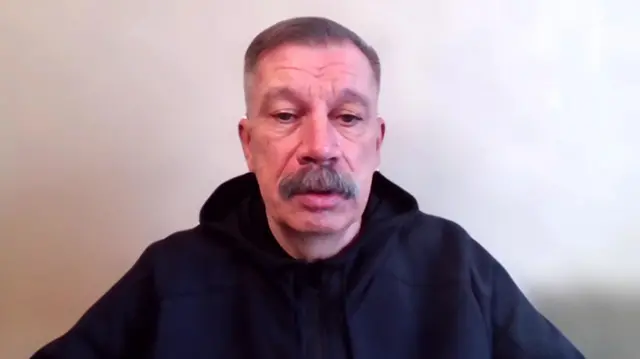 A man in a black hoodie with a moustache looks at the camera