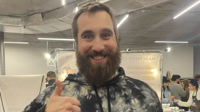 A grinning Maksym Kovalenko wears a tie-dyed hooded jumper and gives a thumbs up in a market.