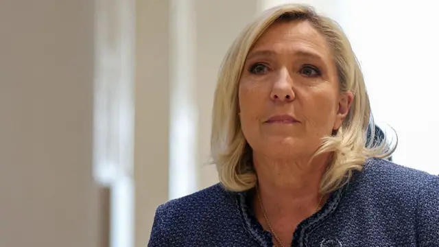 Marine Le Pen in an indigo jacket standing against a beige, well-lit background.