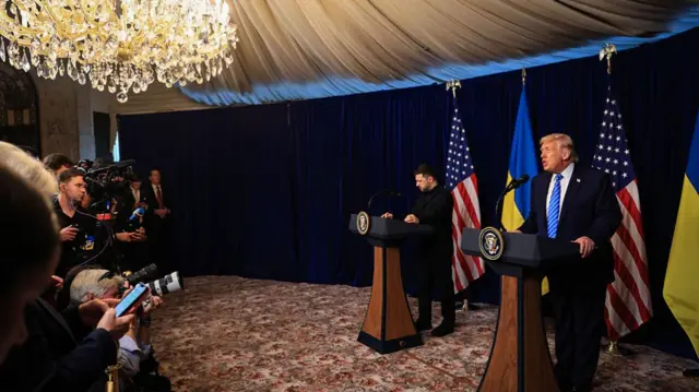 zoomed out view of Trump and Zelensky standing at podiums under a chandelier as reporters ask questions