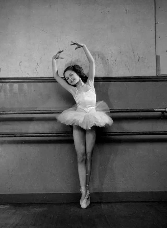 A young Brigitte Bardot in a tutu and ballet slippers, performing a ballet move while standing in front of a wall.