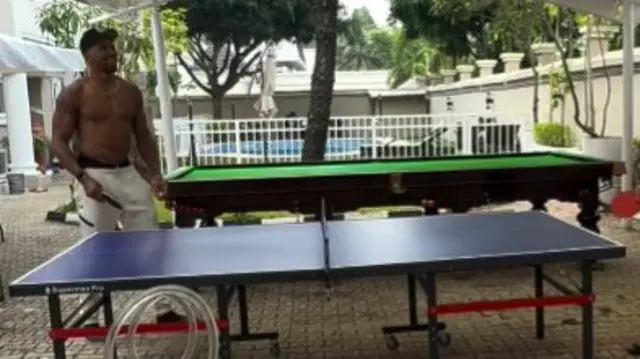 Anthony Joshua standing topless at a table tennis table shirtless holding a paddle