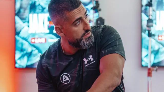 A man in a black t-shirt is looking to the side and there are bright blue posters in the background.