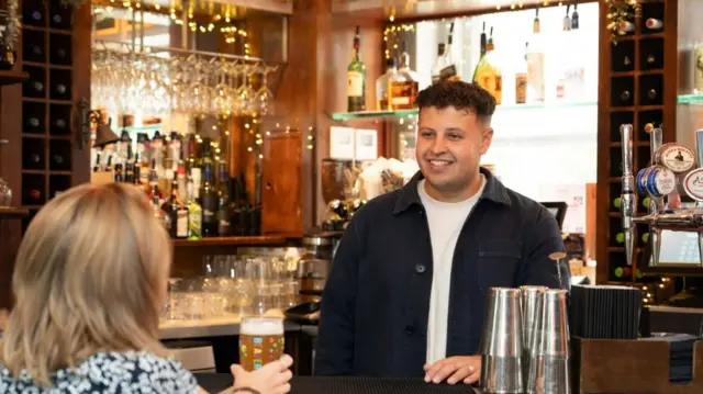 A man stands behind a bar, with a woman with blonde hair sitting at the bar, holding a beer.