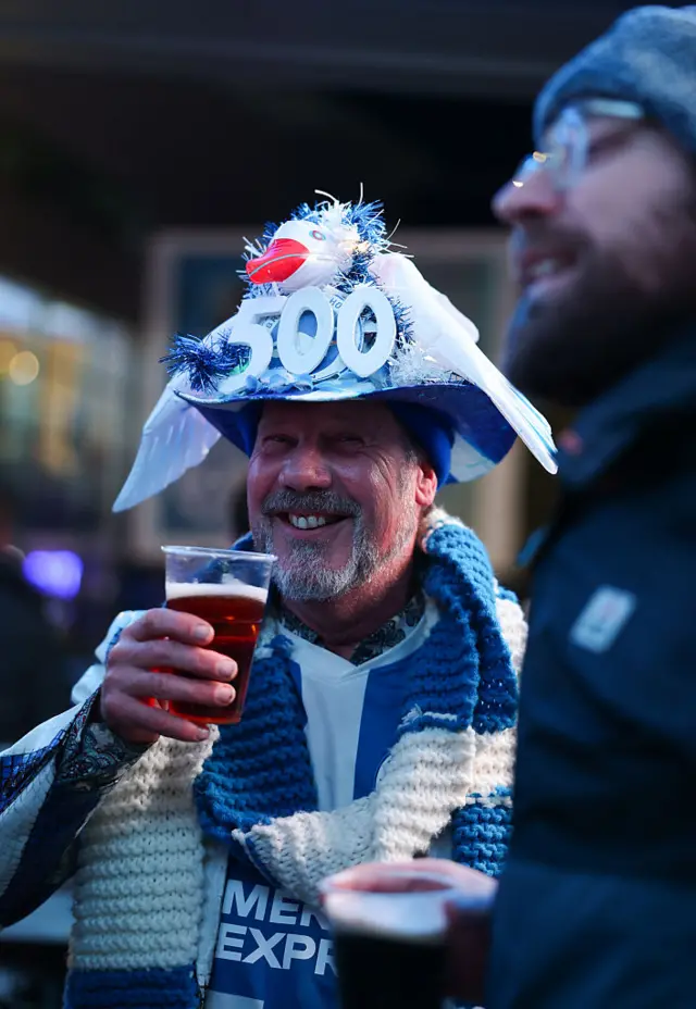 A fan wearing a hat with '500' and a seagull on it, drinking a beer