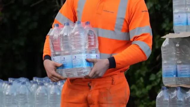 A person in high-vis carrying a case of water. Piles of water bottles can be seen in the background.