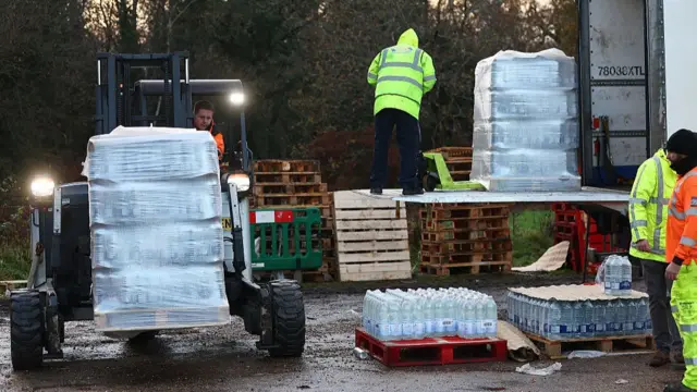 A forklift carrying a pallet of bottled water. men in high-vis clothing are unloading pallets from an open lorry to the right.