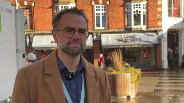 A man in a brown jacket, looking to the side of the frame. There is a brick building with Caffe Nero branding behind him.