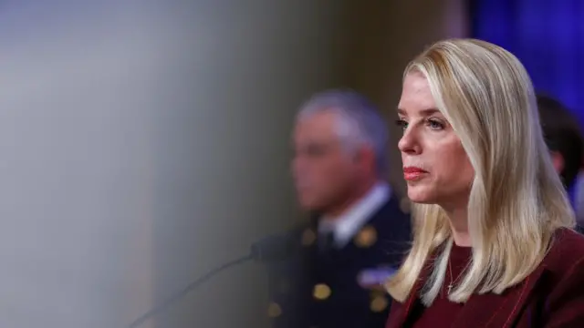 A side-on view of US Attorney General Pam Bondi in front of a lectern, with a blurred out man in the background.