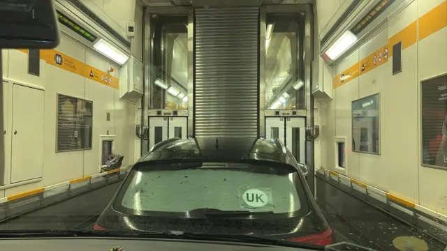 The inside of a LeShuttle train, taken from inside a car with a black car in front and white metal walls of the train surrounding.