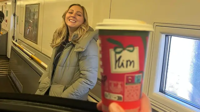 Kim stands smiling in a train carriage behind a takeaway coffee cup being held in front of her with her name on it