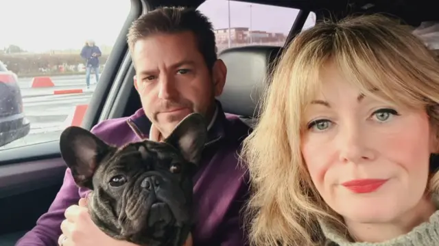 A blonde woman and brunette man sit in a car with a bulldog