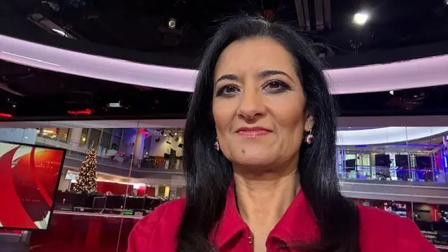 A woman with long dark hair and a red top sits in a studio