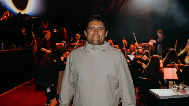 Man in grey jacket smiles in front of an orchestra preparing to play instruments