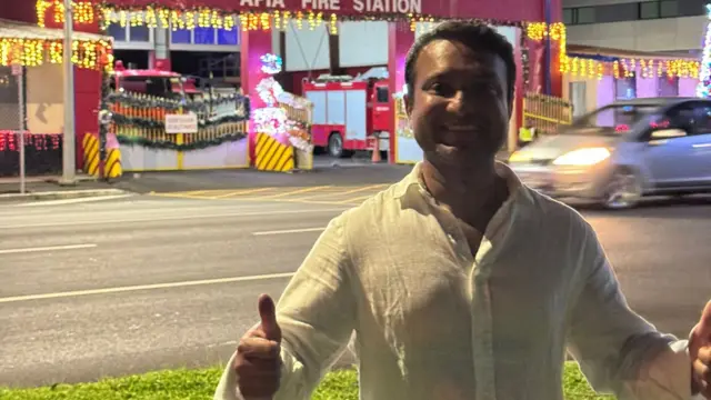 A man stands in front of a fire station decorated with new year lights. He's wearing a linen shirt and holding his thumb's up.