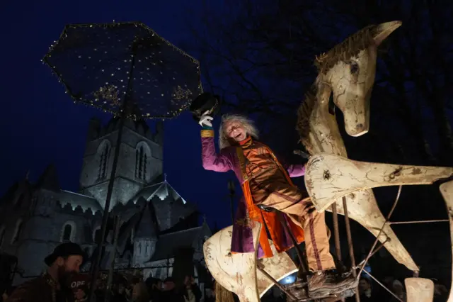A street performer sits on a wooden mechanical horse, smiling and tipping his hat