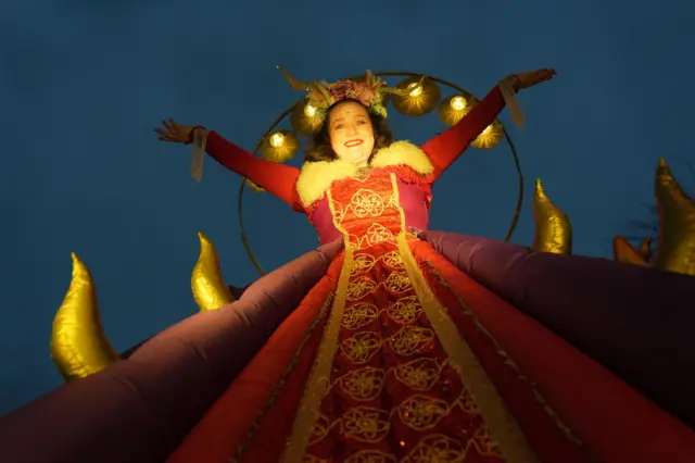 A street performer holds arms a loft, smiling, while wearing an extravagant dress