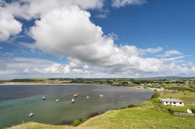 Fishing boats in Chatham Islands in water, surrounded by scattered houses and a cloudy sky