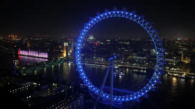 Crowds seen gathering on the banks of the Thames behind the London Eye, which is lit up blue.