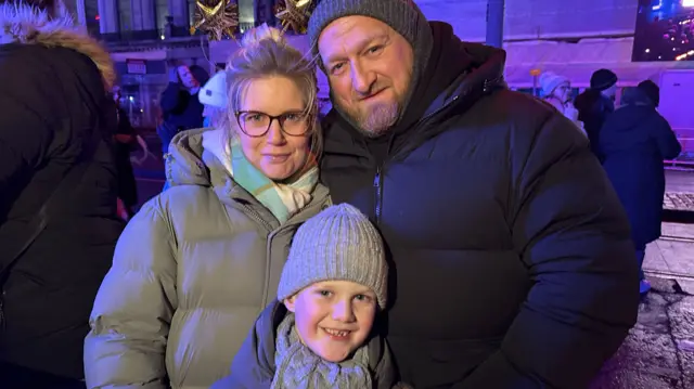 A family, woman man and boy dressed in winter clothes with a street scene behind