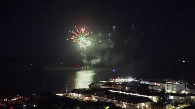 A firework goes off across the water. A number of houses can be seen lit up in the front of the photo