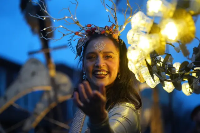 Woman wearing headband to look like antlers looks into the camera and smiles, with her left arm raised forward and pointing towards the camera