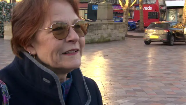 A woman with short red hair in a navy fleece and glasses. She is standing outside and looking at the interviewer.