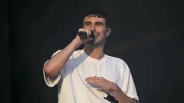 Fred Again speaks into a microphone while performing at Glastonbury Festival, wearing a white t-shirt.