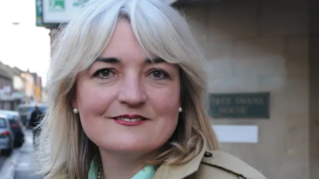 A woman with light blonde hair smiles. She is outside a building, wearing a beige coat.
