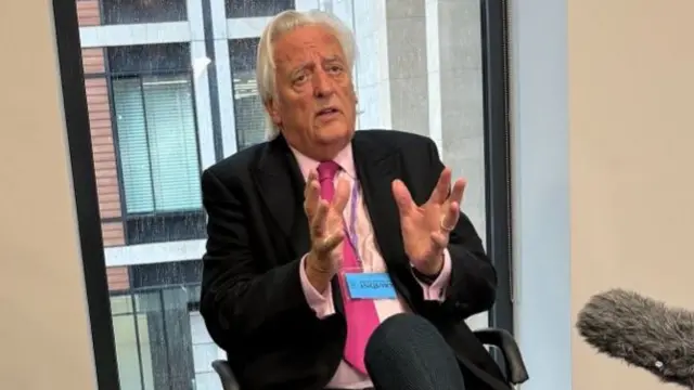 Michael Mansfield KC, who has white hair swept back from his face, gestures with his hands as he talks to the media. He is wearing a black suit and a pink shirt with a pink tie.