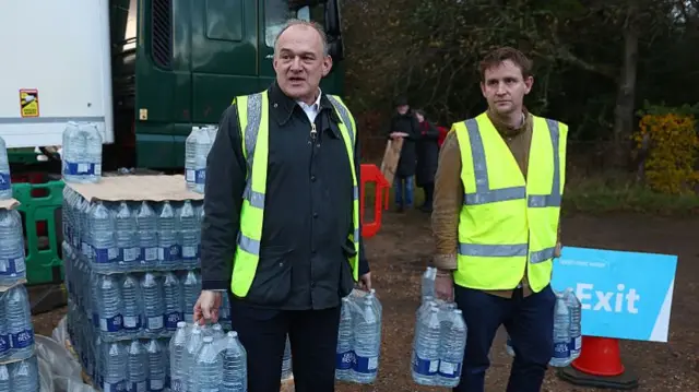Sir Ed Davey MP and Mike Martin MP wearing high-vis jackets and carrying cases of bottled water. A pallet stacked with water and a blue exit sign can be seen behind them.