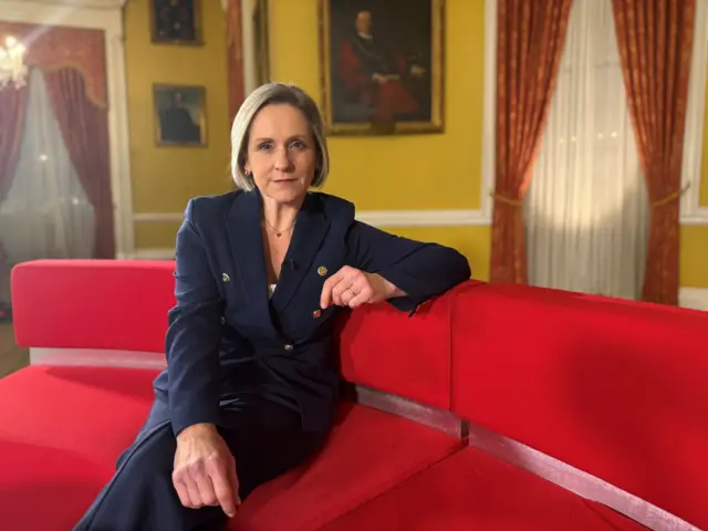 Amanda Parr in a Navy trouser suit sitting on a red sofa. She has one arm on her thigh and one resting on the back of the sofa. There are yellow walls and red curtains behind her.