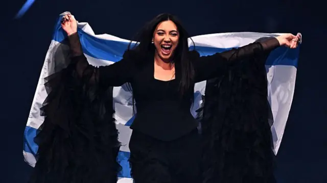 Yuval Raphael, a young, female singer wearing an all-black, frilly dress with long sleeves - is holding an Israeli flag and smiling into the crowd