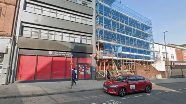 Exterior of Salvation Army in Derby. It is a dark-framed building with red covered windows. A heavily-scaffolded building is to the right.
