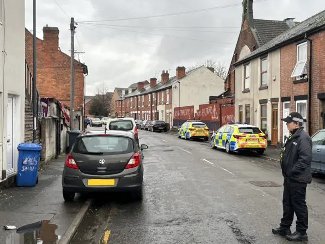 A quiet street with a dark grey car parked on the left pavement and two police cars parked on the right. A uniformed police officer stands in the foreground.