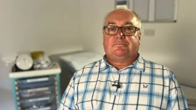 Stephen Cockroft wearing a blue and white plaid shirt and black rectangular framed glasses. He is sitting in a hospital suite with a trolley of medical equipment behind him.