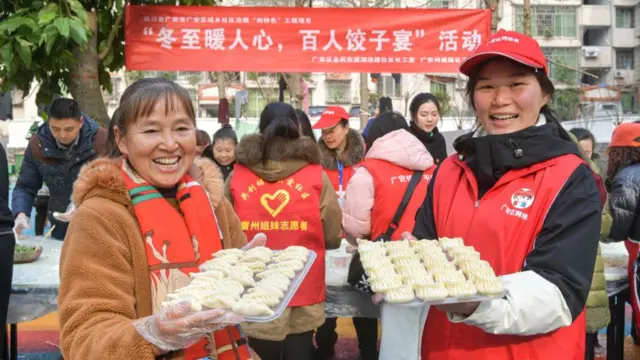 Two women smile at the camera as they hold up trays with dumplings on them
