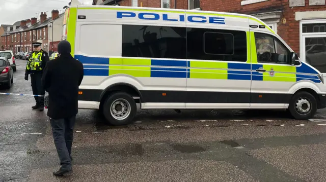 A police van parked across a residential street with a uniformed police officer stood next to it.
