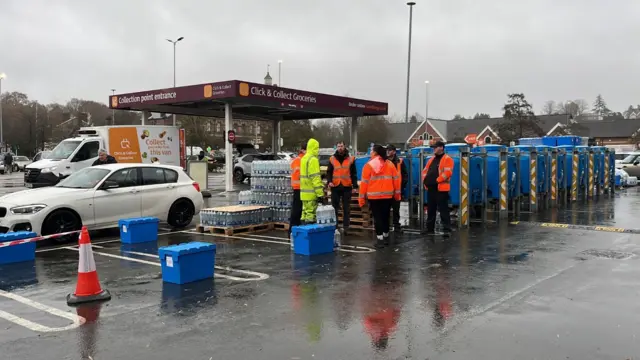 People in high-vis clothing stood beside pallets of bottled water and large blue tanks. Cars and a van are seen in the car park behind them.