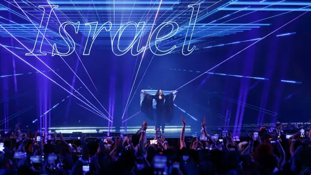 Yuval Raphael holding an Israeli flag behind her in front of the Eurovision audience. The word 'Israel' is lit up behind her on the stage
