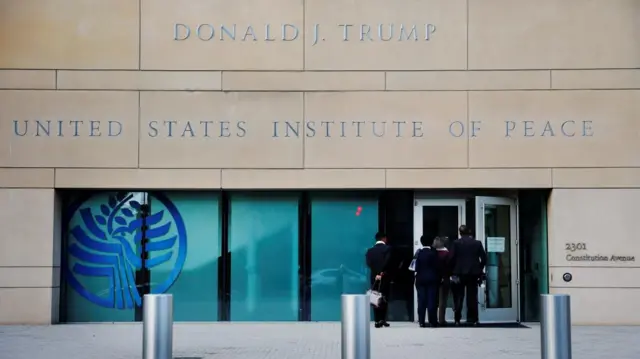 A sign reads 'Donald J. Trump' on a stone building. The words 'United States Institute of Peace' are in block capitals underneath and several people can be seen entering the building