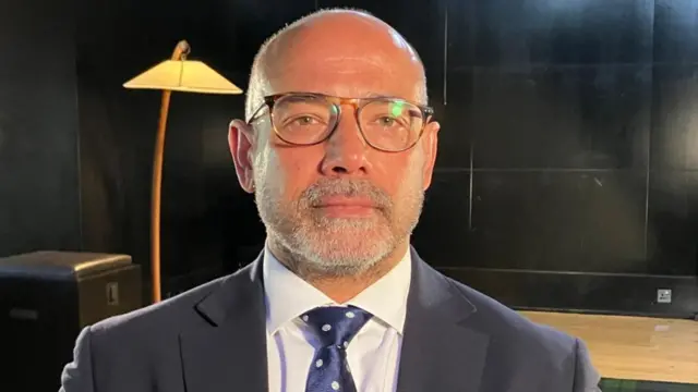 A head and shoulders image of Neil Basu wearing a dark suit, white-collared shirt and blue polka dot tie. He is sitting in a black studio with a mid-century modern lamp behind him.