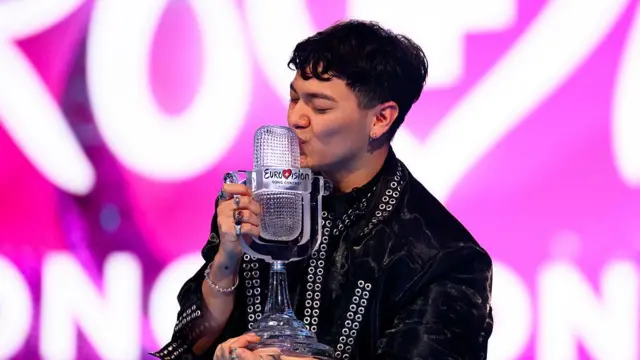 JJ representing Austria celebrates after winning Eurovision 2025. He is kissing the trophy, a translucent retro microphone, and wearing a shiny black outfit.