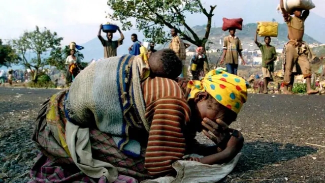 A Rwandan woman collapses with a child on her back alongside the road connecting Kibumba refugee camp and Goma in 1994. Several people carry supplies on their heads in the background.